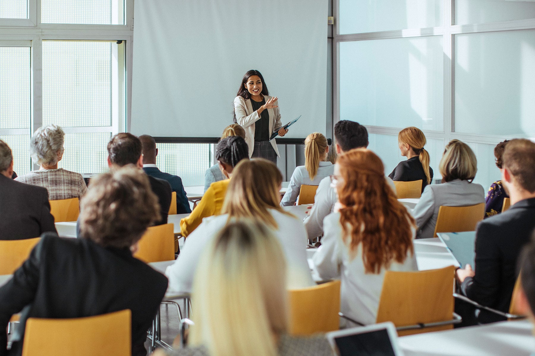 A woman in her 30s in front of a whiteboard speaking to a group of 20 students in a classroom